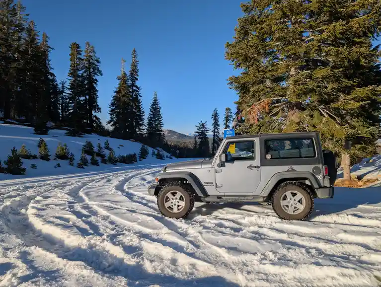 A silver Jeep Wrangler is parked on a snowy, winding road surrounded by tall evergreen trees under a clear blue sky. A blue…