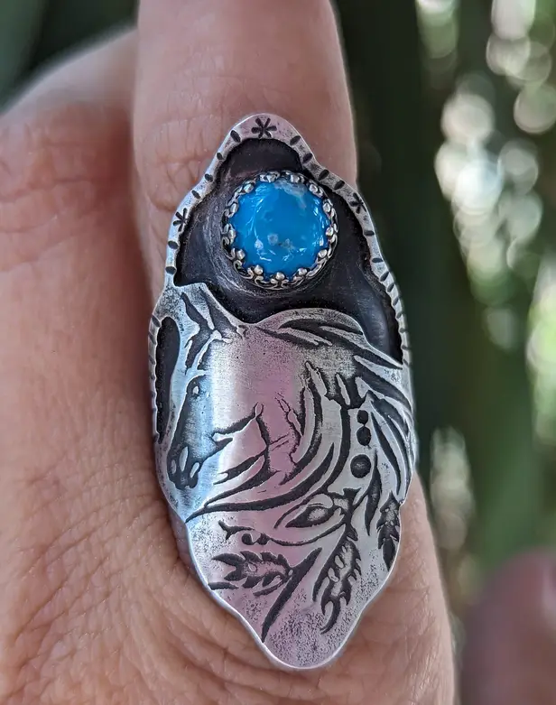 A close-up photo shows a large, ornate silver ring on a finger, featuring an engraved horse head design and a prominent blue…