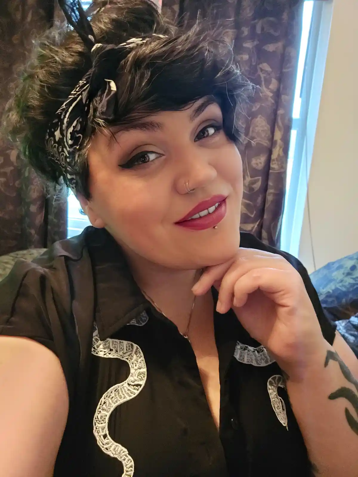 A person with short dark hair, wearing a black bandana, a black shirt with white snake embroidery, and a snake tattoo on…