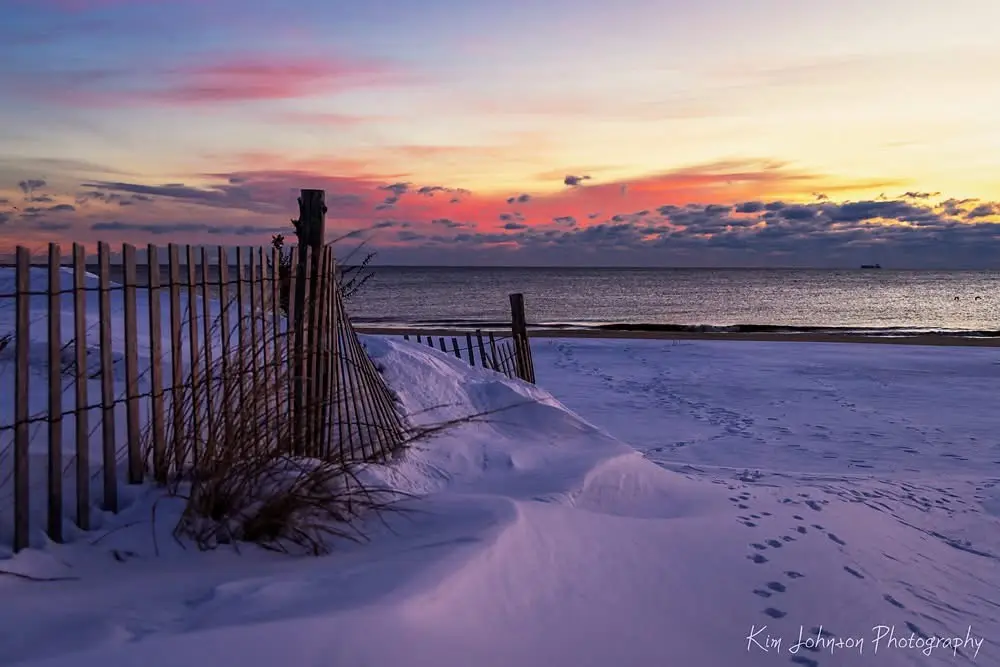 A serene winter beach scene at sunset, with snow blanketing the sand and a wooden fence in the foreground. The sky displays…