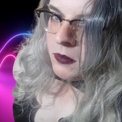 A person with silver-gray wavy hair, wearing glasses and dark lipstick, looks directly at the camera against a dark…