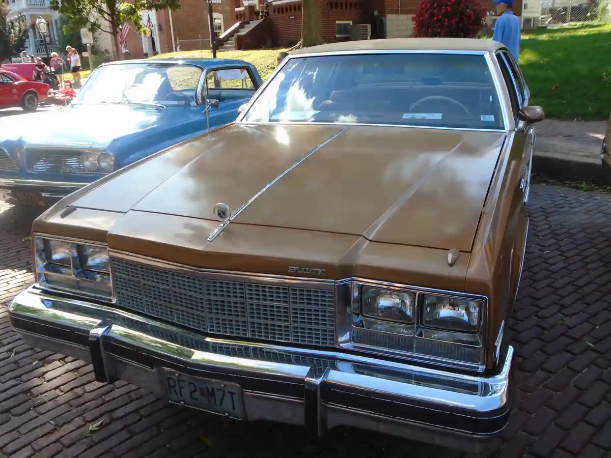 A front-facing photo of a brown 1970s Buick sedan parked on a brick road at a car show, with other vintage cars and people…