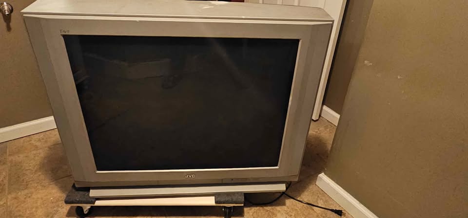 A large, old JVC CRT television with a thick silver frame sits on a wheeled stand in a room with beige walls and tiled…
