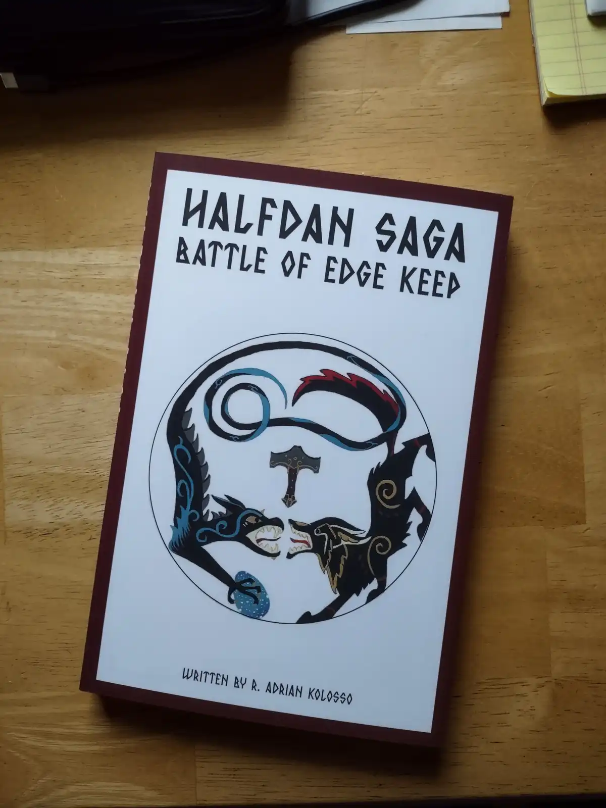 A book titled 'HALFDAN SAGA BATTLE OF EDGE KEEP' by R. Adrian Kolosso sits on a wooden desk, featuring a circular…