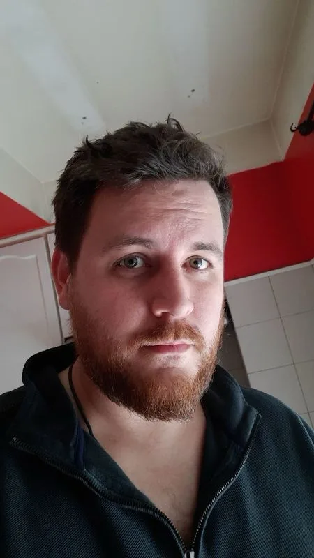 A man with a reddish beard and green eyes stares directly at the camera with a deadpan, unimpressed expression. He is…