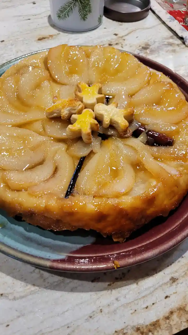 A close-up photo of a baked pear tart on a ceramic plate, featuring glossy sliced pears arranged in a circular pattern, with…