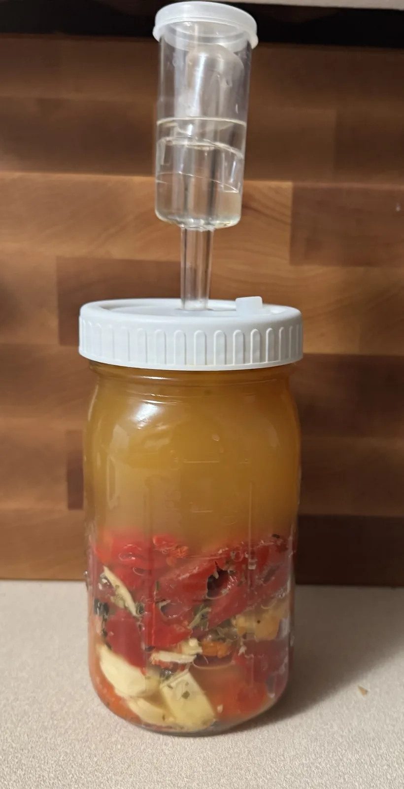 A glass mason jar filled with chopped red tomatoes, garlic, and herbs submerged in a cloudy orange liquid, topped with a…