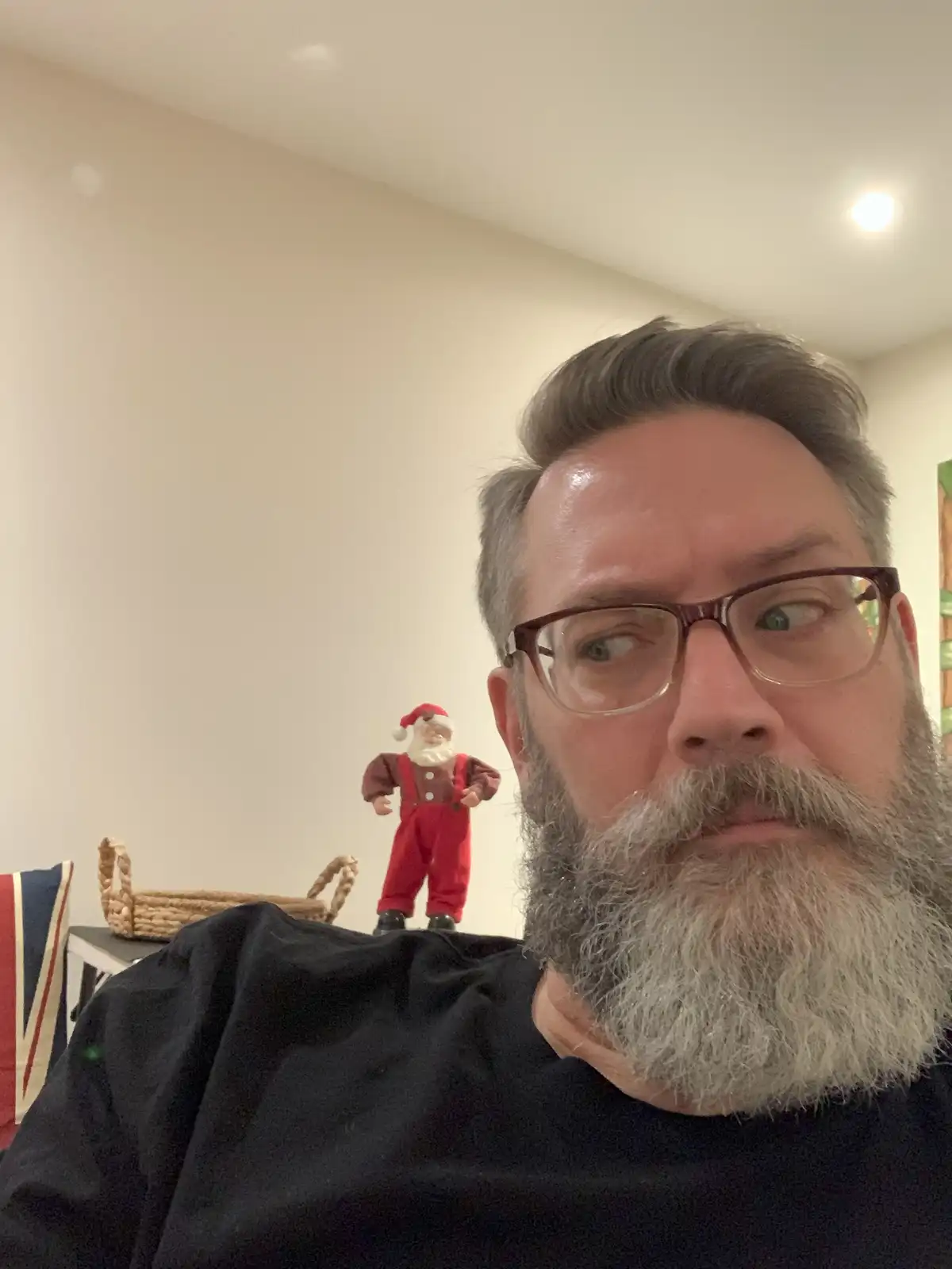 A man with glasses and a gray beard looks over his shoulder with a startled expression, as a small Santa Claus figurine…