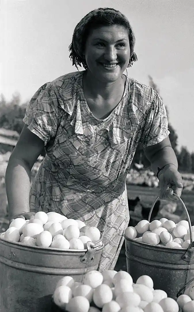 A black-and-white photo shows a smiling woman outdoors holding two buckets overflowing with eggs, with more eggs piled in…