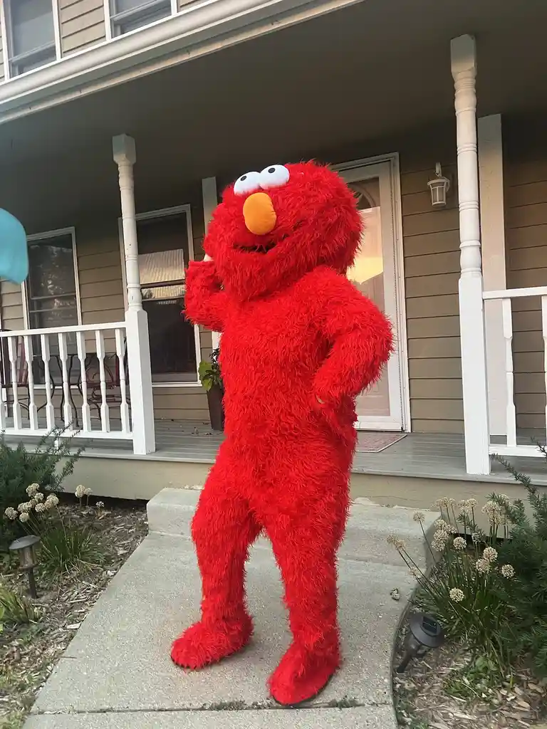 A person in a full-body, fluffy red Elmo costume stands on a concrete walkway in front of a house with a porch. The…
