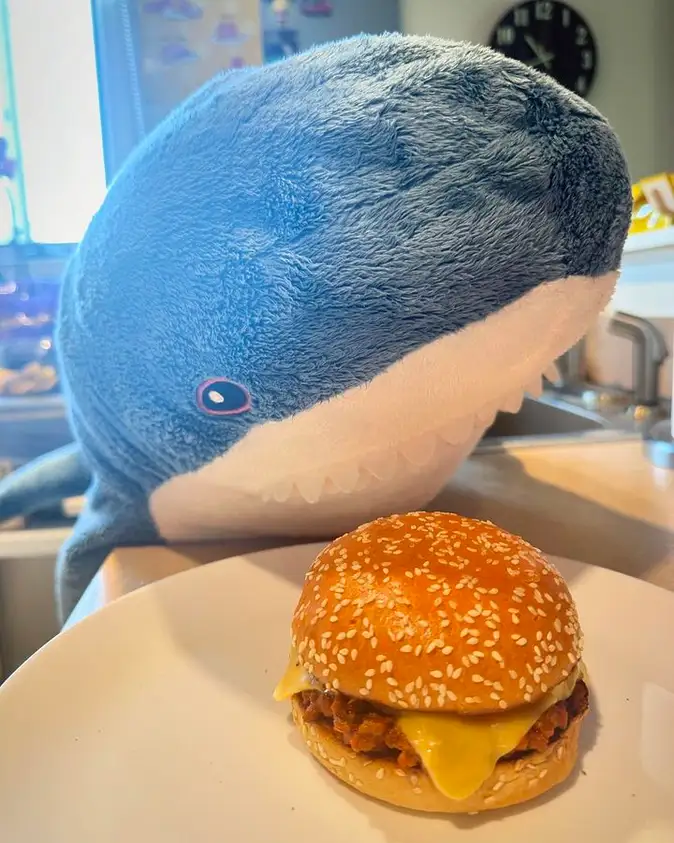 A large blue plush shark toy with a white underbelly and visible teeth leans over a white plate holding a sesame seed burger…