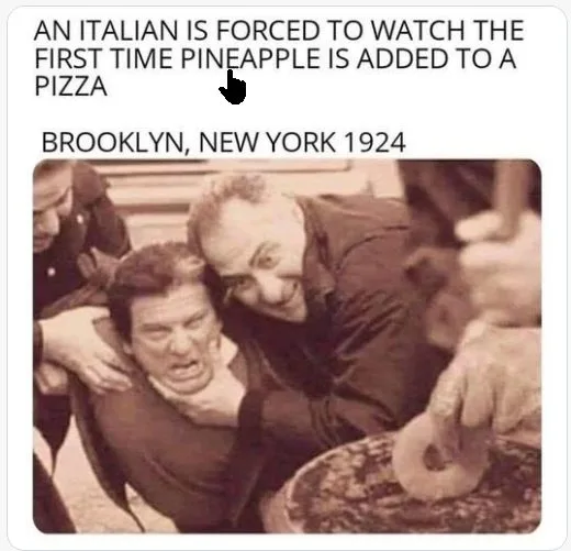 A sepia-toned photo shows a man being held by another man who is grinning while holding a pineapple slice over a pizza. The…