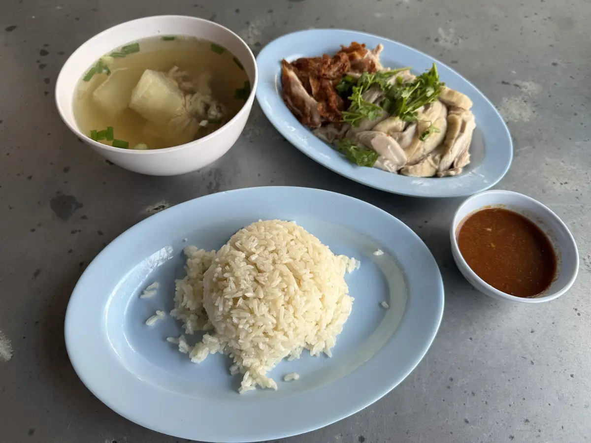 A meal featuring a mound of steamed rice on a light blue plate, a plate of sliced chicken with herbs, a bowl of clear soup…