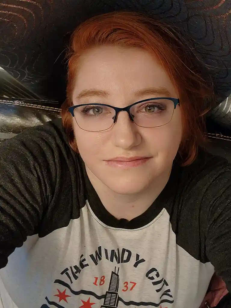 A person with red hair and glasses wearing a 'The Windy City' t-shirt lies on a patterned couch, looking directly at the…