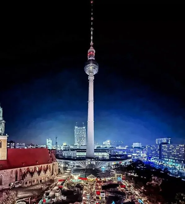 A nighttime photo of the Berlin TV Tower (Fernsehturm) illuminated against a dark sky, with a festive market and city lights…