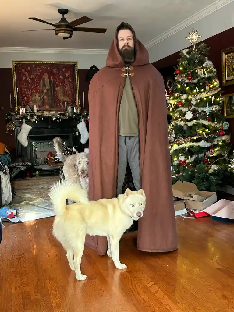 A man with a beard stands in a living room decorated for Christmas, wearing a large brown hooded cape that covers his entire…