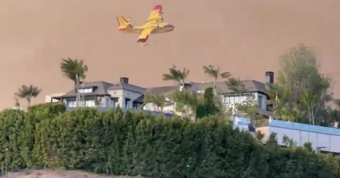 a yellow firefighting aircraft flies over a residential area with houses and palm trees under a hazy, orange-brown sky. The…