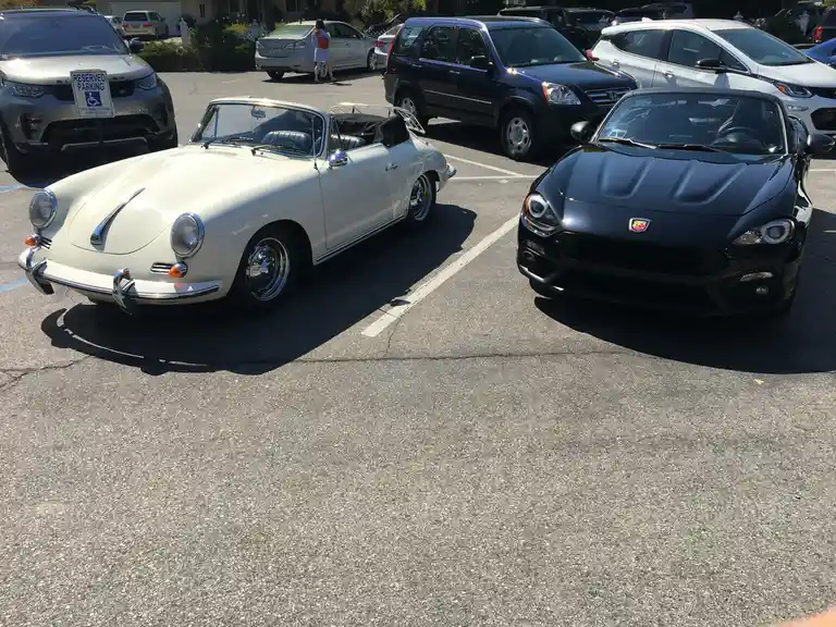 Two cars are parked side-by-side in a lot: a classic white Porsche 356 convertible and a modern black Fiat 124 Spider. A…