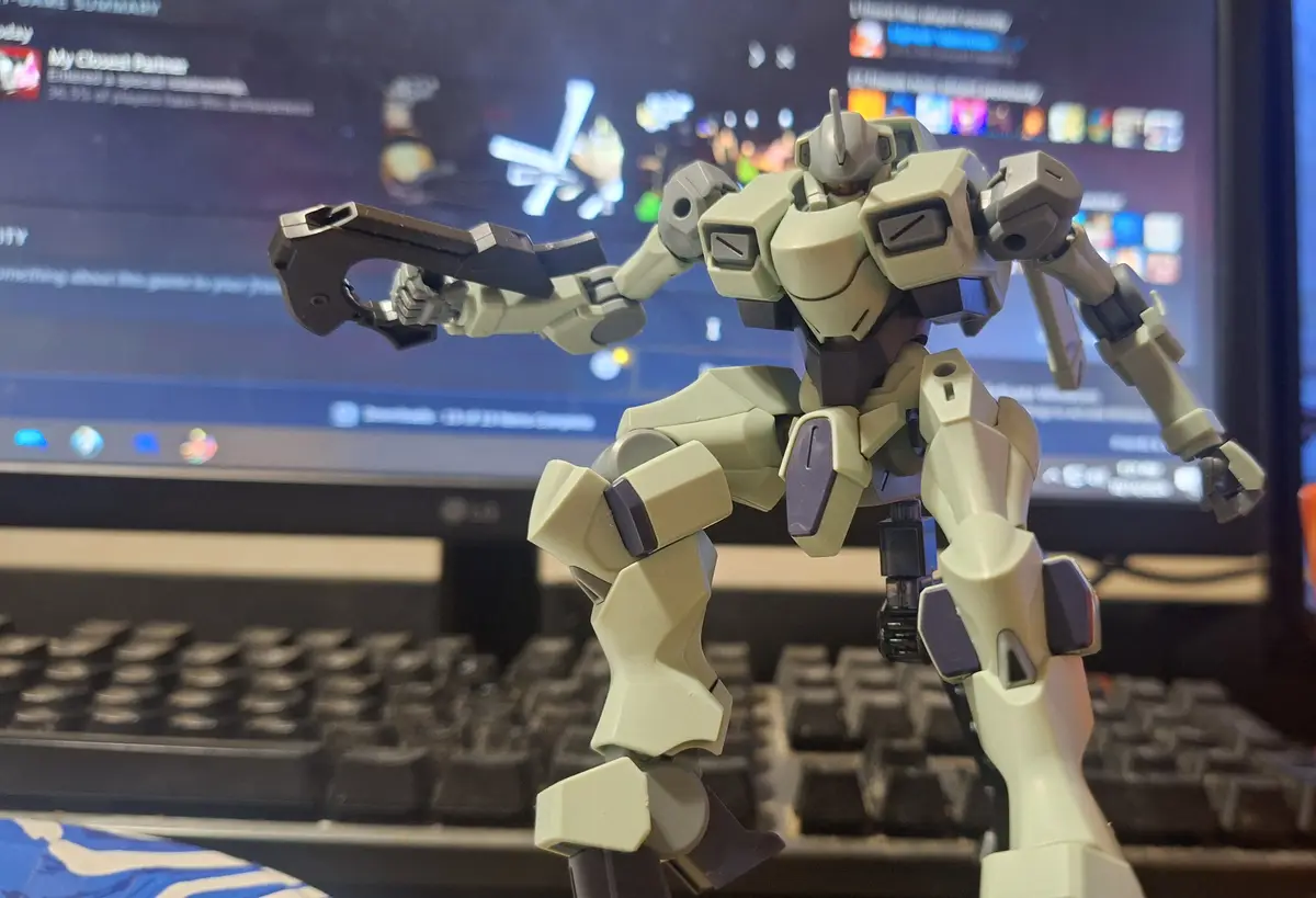 A beige and gray plastic robot model, resembling a mecha from an anime, stands in front of a computer monitor displaying a…