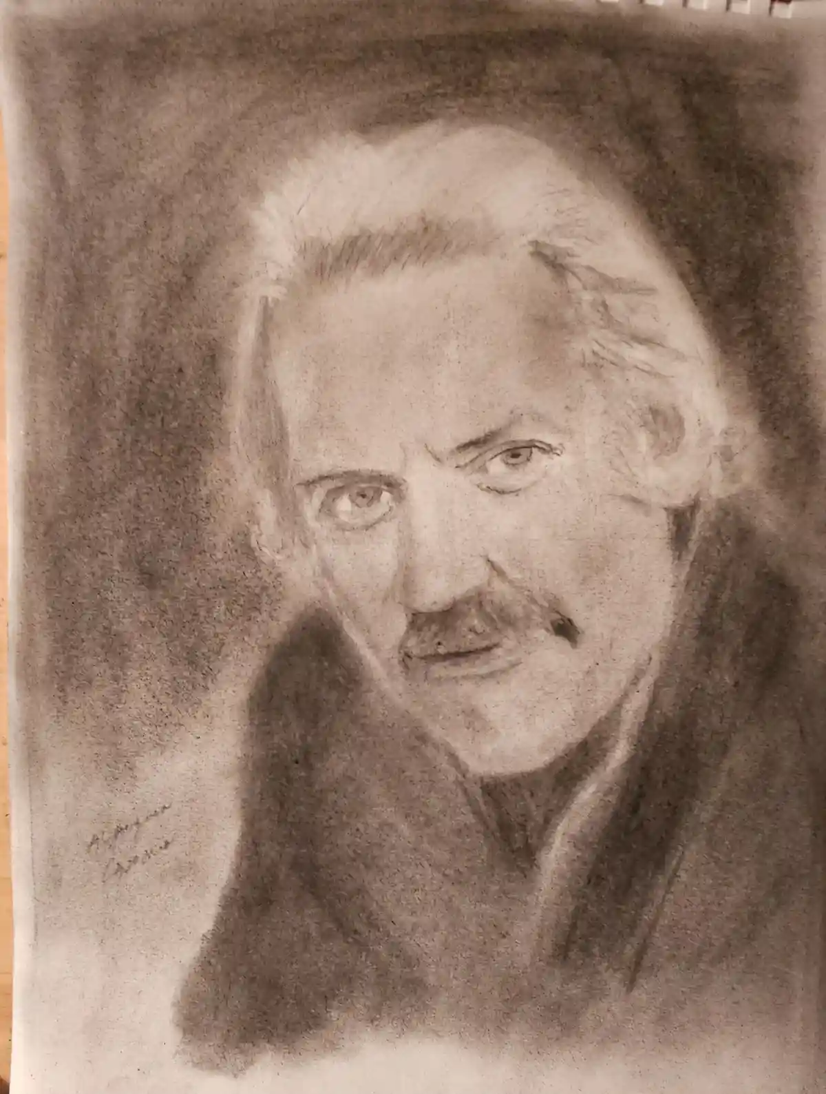 A detailed charcoal sketch of a man with a mustache and intense gaze, drawn on paper with a spiral notebook binding visible…