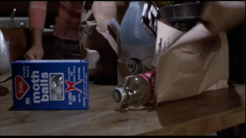 A blue box of Eno's Pure Pana moth balls sits on a wooden table next to a spilled soda bottle and a paper bag, with a…
