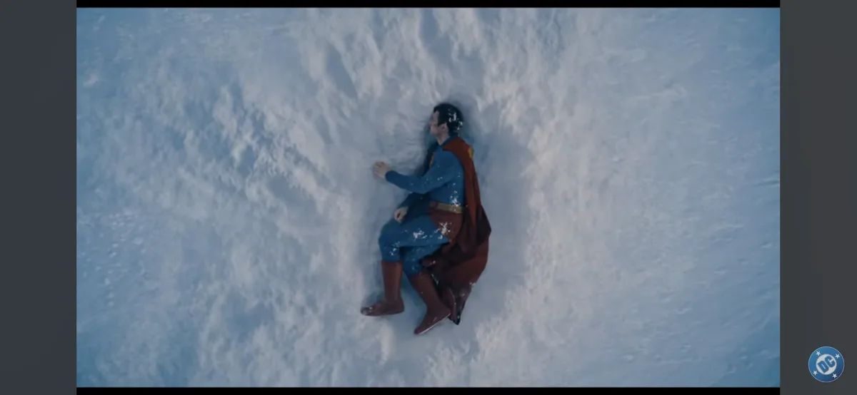 A man in a blue and red superhero costume with a cape is sitting in a vast snowy landscape, partially buried in snow up to…