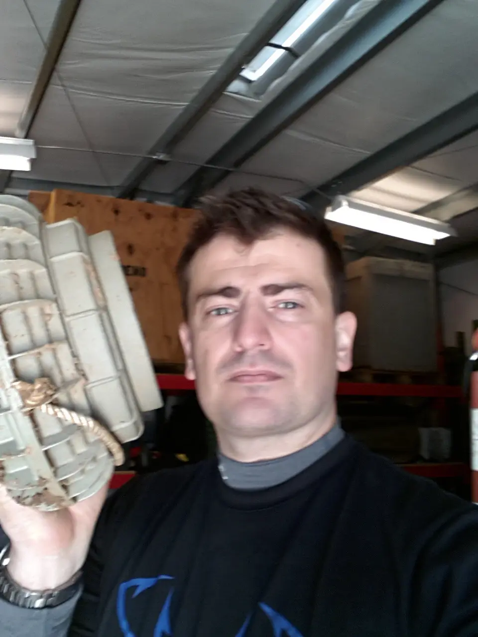 A man in a garage holds up a large, rusty, segmented metal object resembling a grenade or bomb casing, looking confused and…