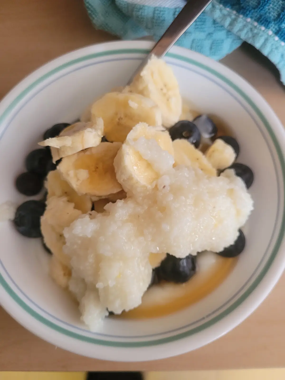 A close-up photo shows a white bowl with green and blue stripes containing a messy breakfast of sliced bananas, blueberries…