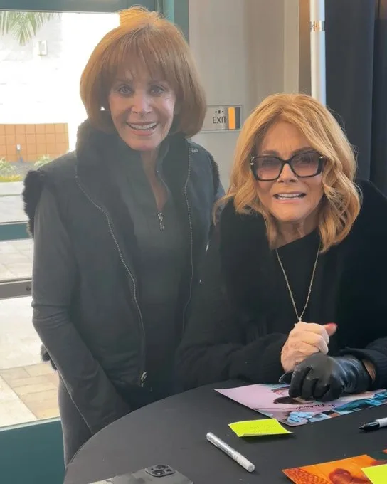 Two women, one standing and one seated at a table with pens and fan items, both smiling. The seated woman wears black gloves…