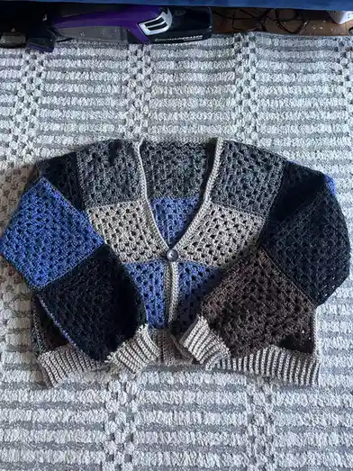 A hand-crocheted, patchwork-style cardigan with blue, black, brown, and beige sections lies flat on a striped rug. A purple…