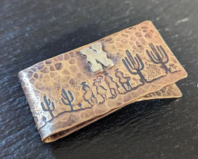 A copper-colored money clip with a hammered texture features an embossed scene of three cowboys on horseback and several…