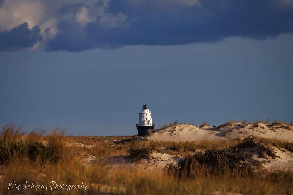A solitary white lighthouse stands atop sandy dunes under a dramatic sky with dark clouds overhead and golden grass in the…