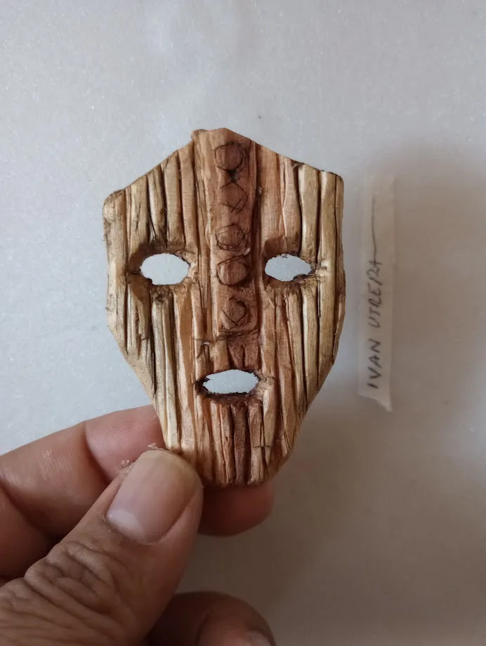 A hand holds a small, crudely carved wooden mask with three oval cutouts for eyes and mouth, resembling a primitive or…