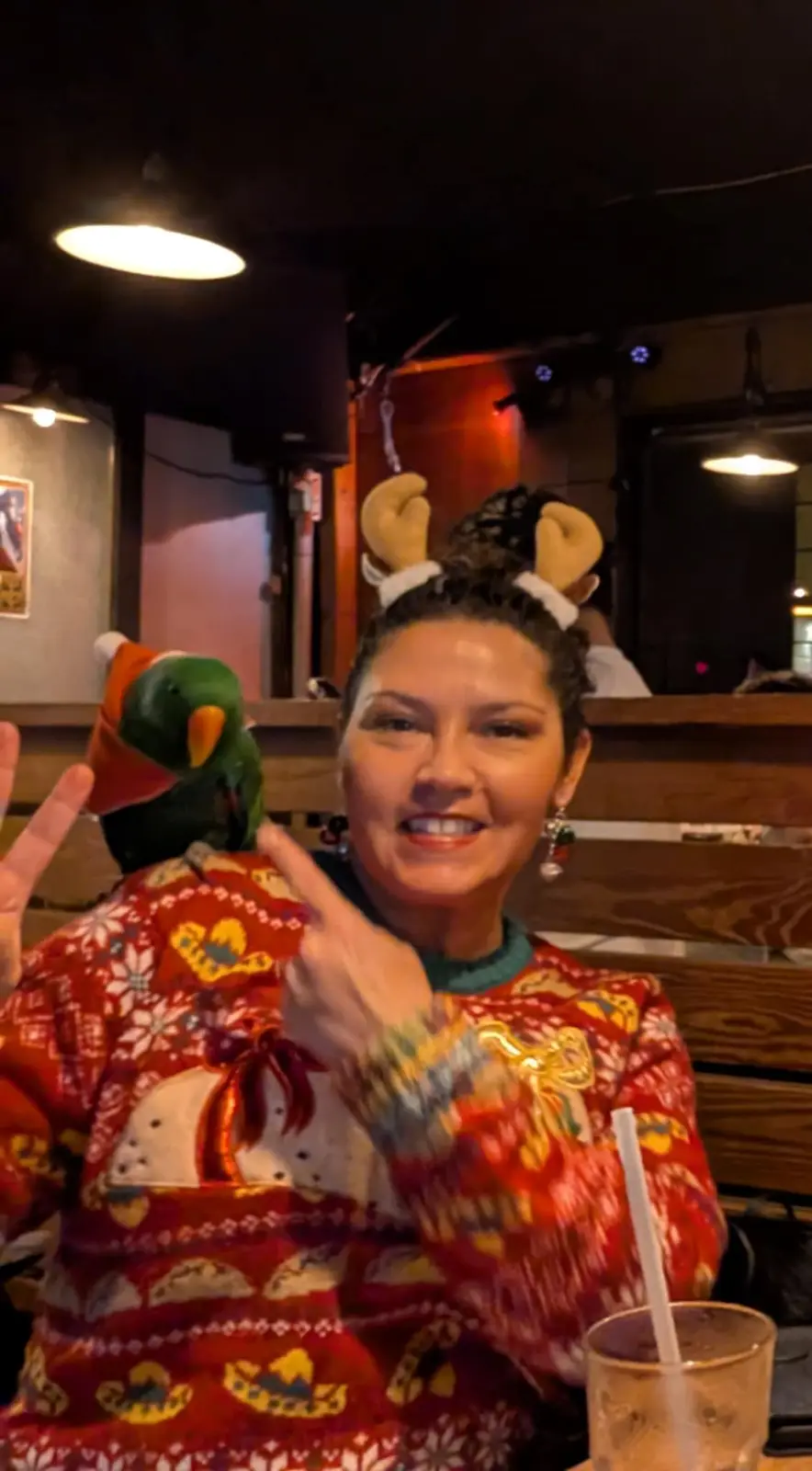 A woman wearing a festive red sweater with taco and snowflake patterns and reindeer antlers sits at a table, smiling and…
