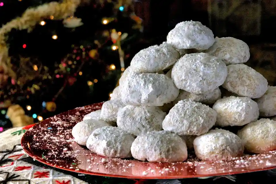 A tall pyramid of powdered sugar-coated snowball cookies sits on a red plate, with a blurred Christmas tree and lights in…