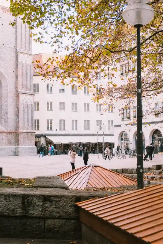 A scenic autumn city square with people walking, trees with yellow leaves, and a brick building in the background. The image…