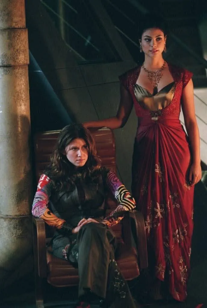 Two women from the movie 'The Hunger Games' are pictured; one sits in a chair looking annoyed while the other stands beside…