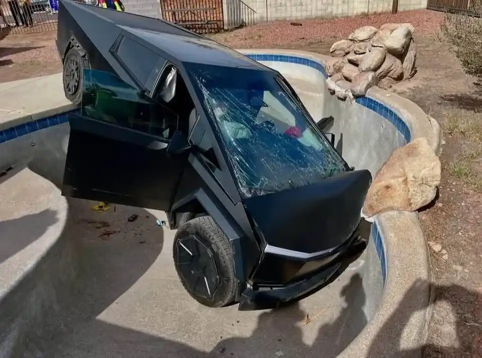 A black, angular Tesla Cybertruck is severely damaged, partially submerged in an empty swimming pool with cracked windshield…