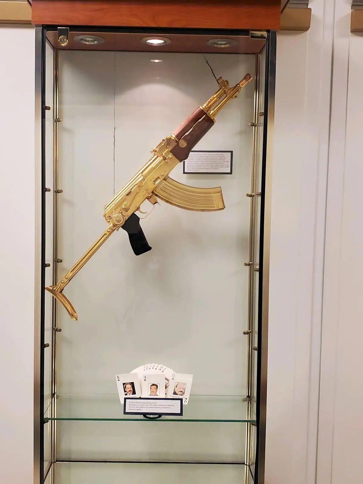 A gold-plated AK-47 rifle is mounted diagonally inside a glass display case, with a small plaque nearby. On the bottom…