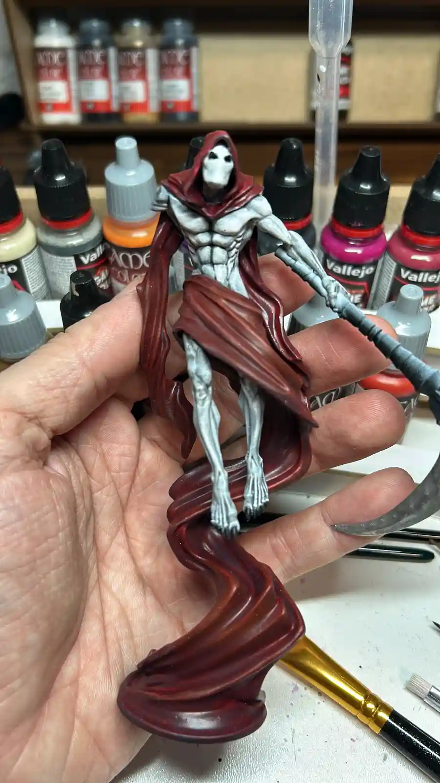 A hand holds a painted miniature of a grim reaper figure with a skeletal face, red hooded cloak, and scythe, surrounded by…