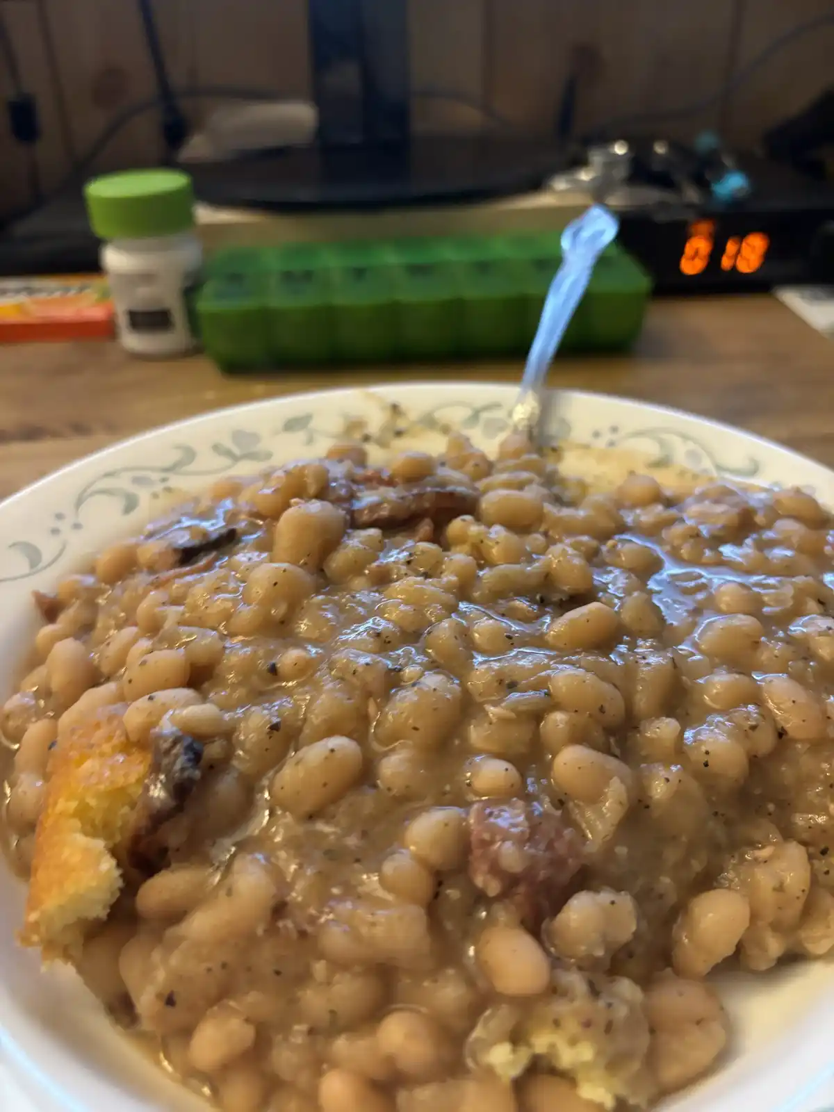 A close-up photo of a white bowl filled with creamy baked beans, mixed with bits of bacon and cornbread, with a spoon stuck…