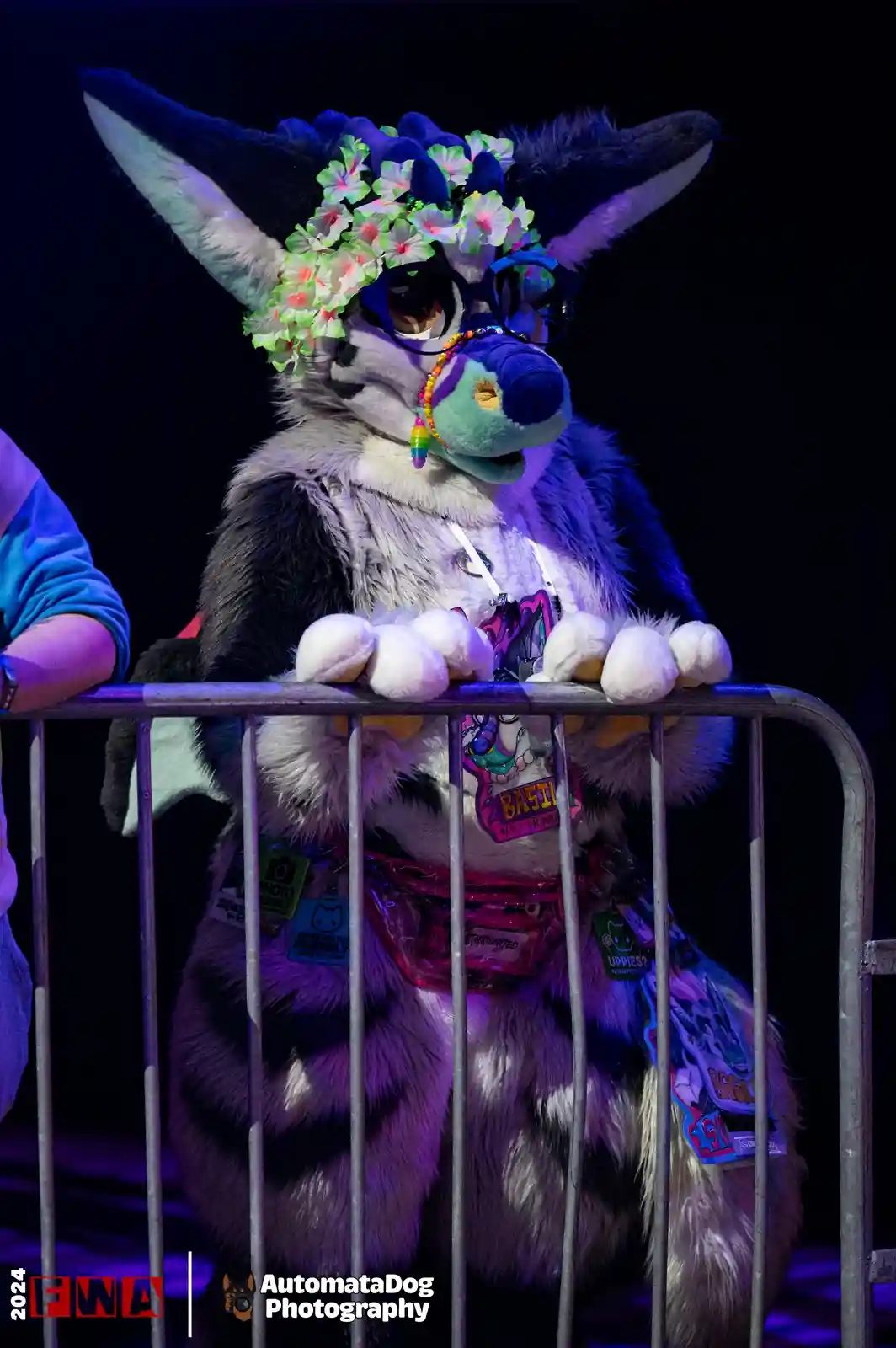A person in a detailed black-and-white fursuit with large ears, a colorful flower crown, and glasses leans over a metal…