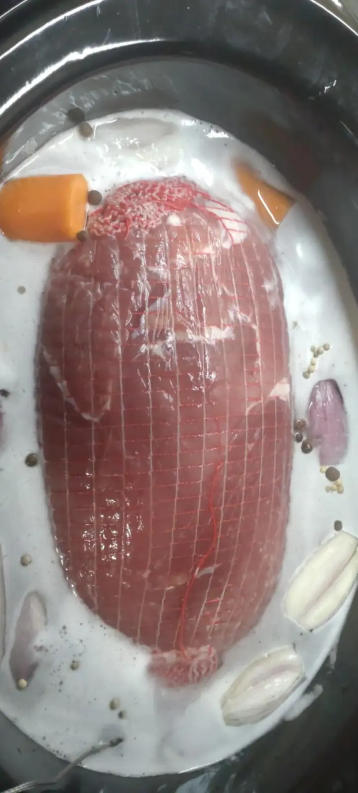 A raw, red meatloaf wrapped in red string sits in a metal bowl submerged in white liquid, surrounded by orange carrot…