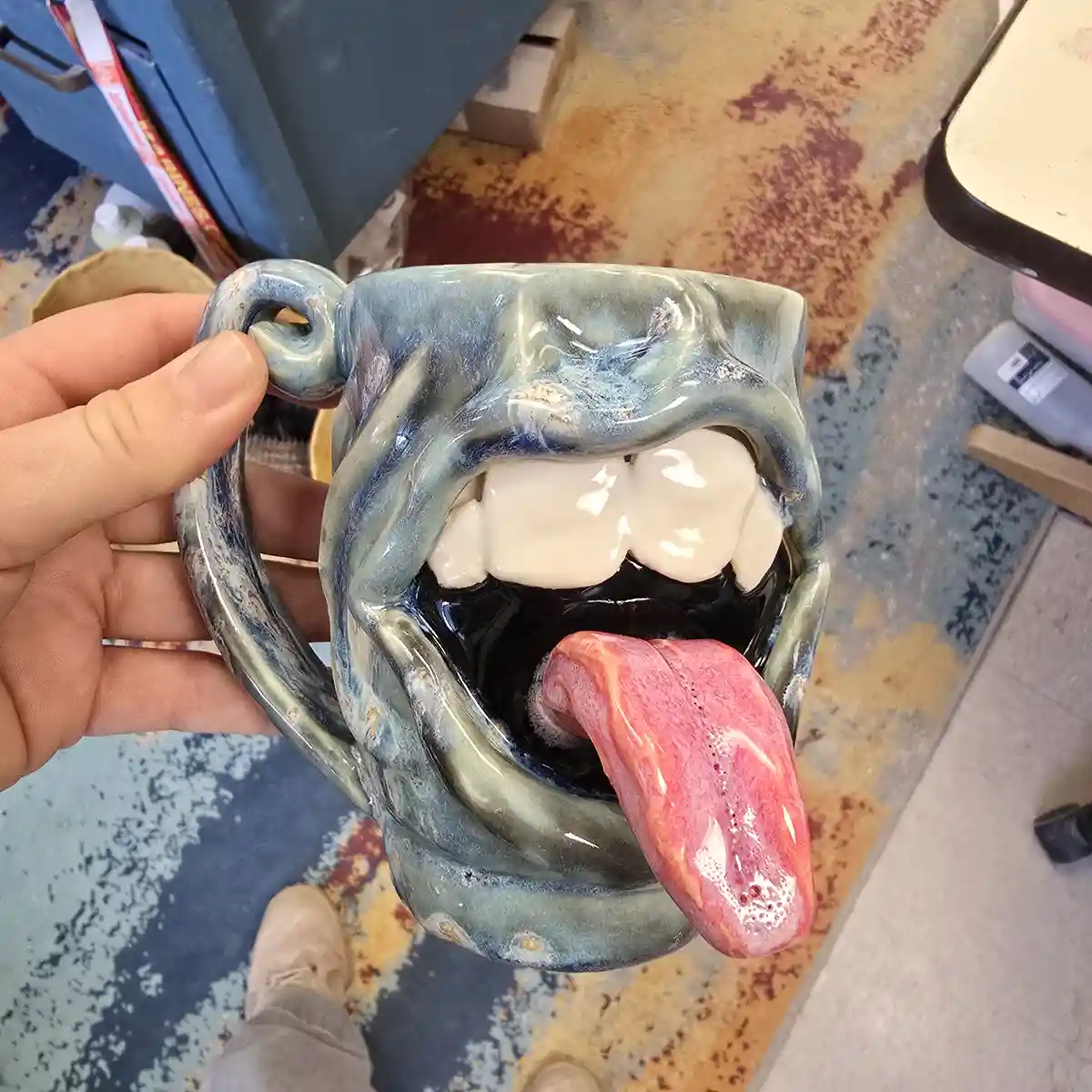 A hand holds a ceramic mug shaped like an open mouth with visible teeth and a long, protruding pink tongue. The mug has a…