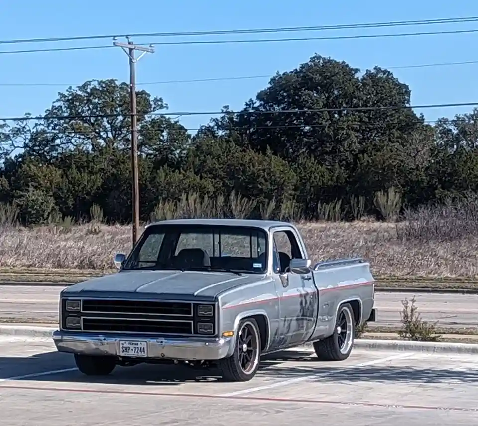 A silver classic Chevrolet pickup truck with black wheels and a red stripe is parked in a lot under a clear blue sky, with…