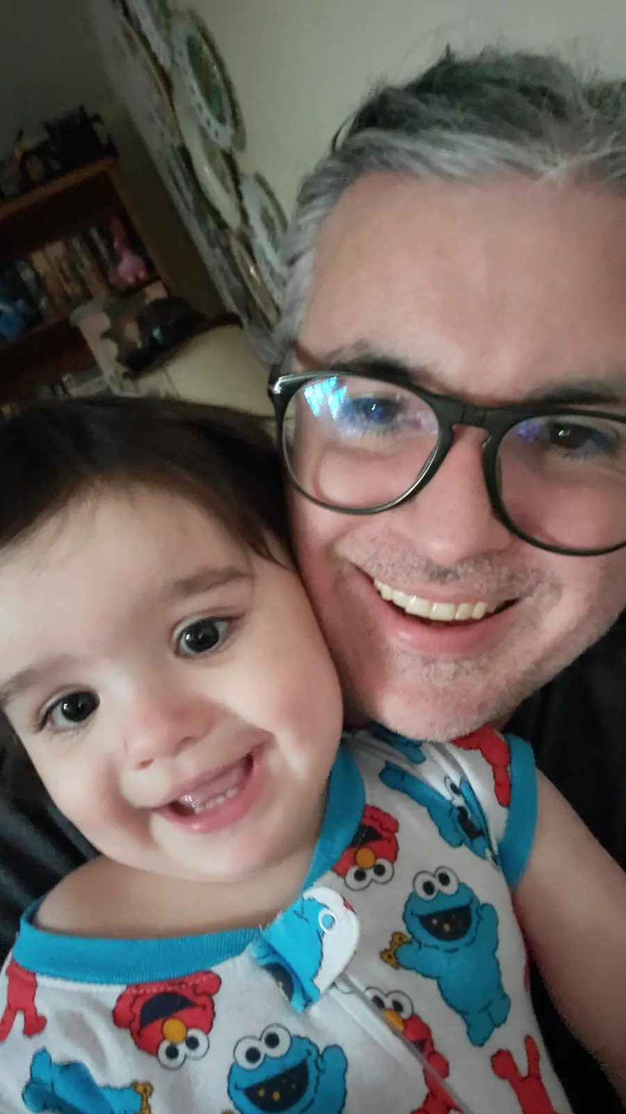 A close-up selfie shows a smiling adult man with glasses and gray hair holding a smiling toddler. The toddler wears a white…