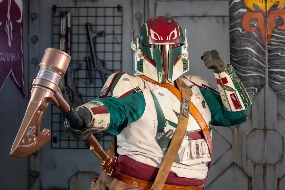 A person in a detailed Boba Fett costume, holding a large hammer-like weapon, poses with one fist raised. The background…