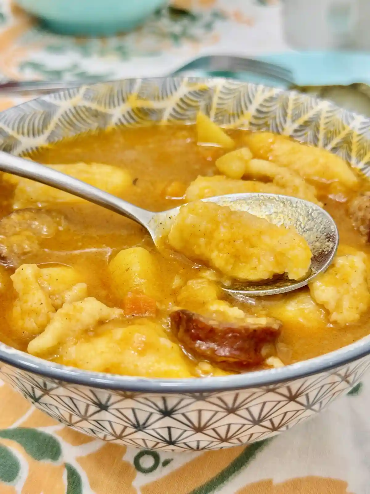 A close-up photo shows a spoon lifting a chunk of soft, pale dumpling from a bowl of thick, orange-colored curry. The bowl…