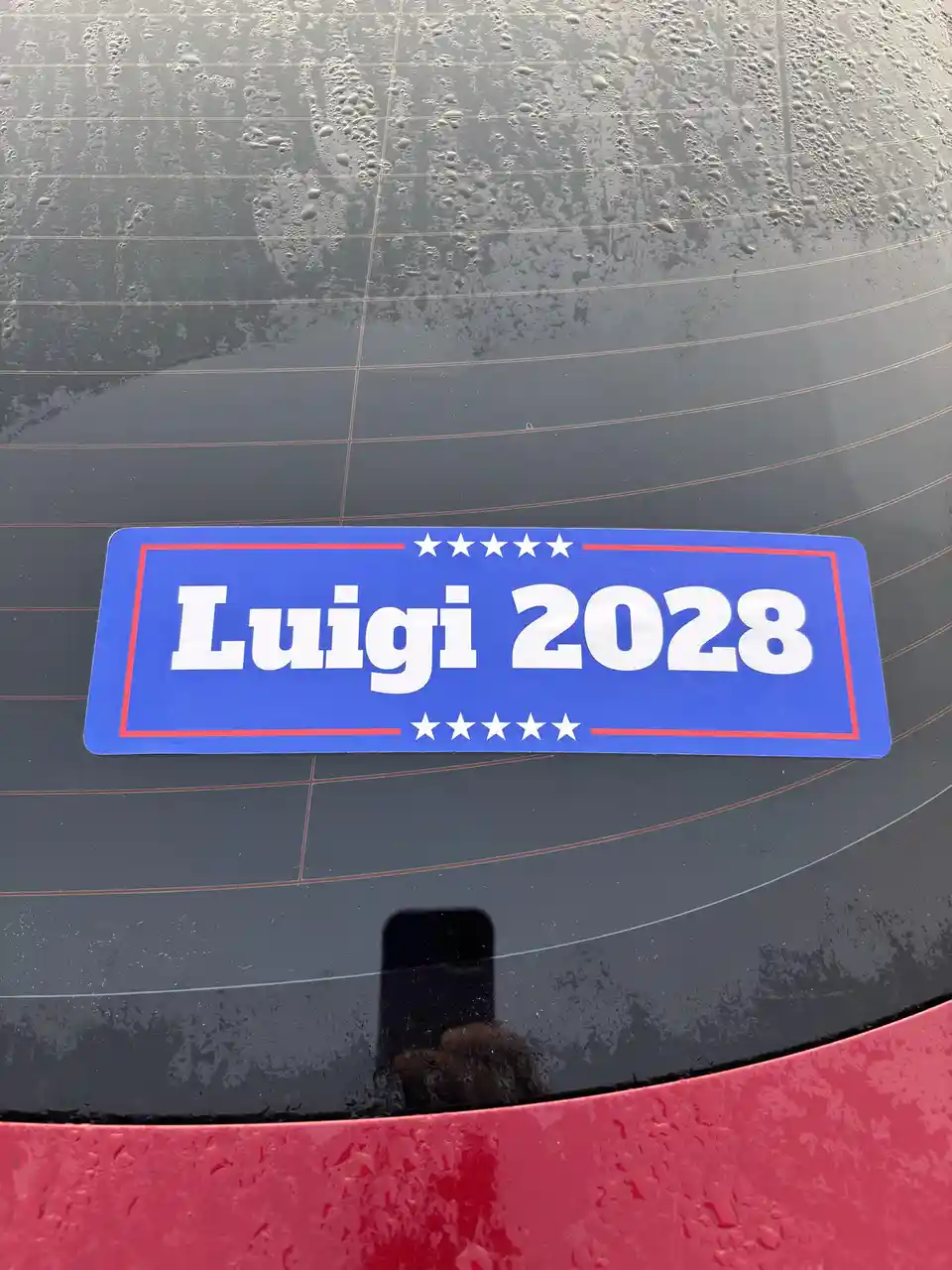 A blue political-style sticker with red borders and white stars reads 'Luigi 2028' and is affixed to the rain-speckled rear…