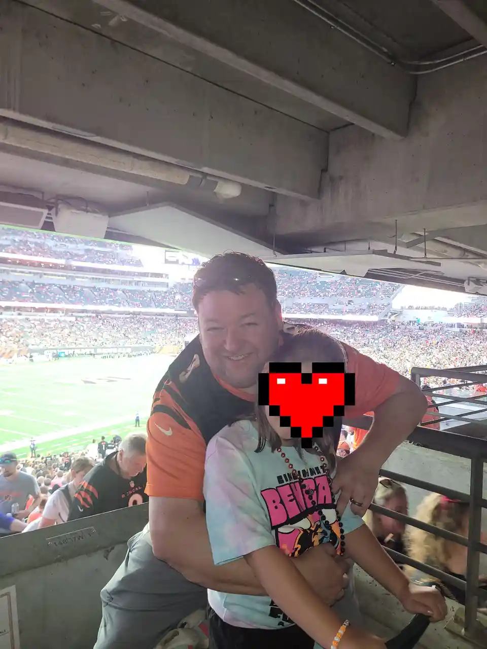 A man in a Cincinnati Bengals jersey hugs a young girl wearing a Bengals-themed shirt at a crowded football stadium; her…